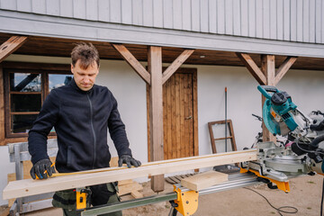Man working outdoors with a miter saw, measuring a wooden board in front of a house, preparing for...