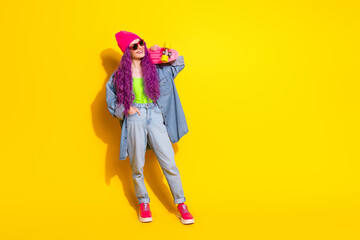 Youthful woman with vibrant pink hair holding a skateboard in front of a bright yellow background wearing colorful and trendy fashion