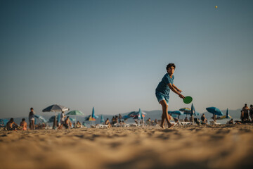 A young man enthusiastically plays paddle ball on a vibrant beach filled with people.