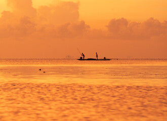 Fototapeta premium “Silhouettes of local fishermen on a traditional wooden boat at sunrise in Thale Noi, Phatthalung, southern Thailand. The peaceful golden morning light reflects off the calm water, capturing the beaut
