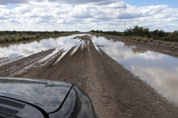 A car parked in front of a long stretch of flooded land that has tire marks from cars that have...