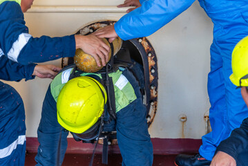 Crew member wearing breathing apparatus exits a narrow hatch after providing first aid during a man evacuation drill from a confined space on a merchant ship.