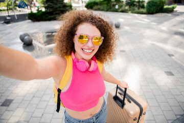 Joyful young woman with curly hair and pink top takes a selfie outdoors while traveling with her suitcase, embracing summer vibes