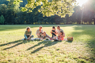 Big family sitting on picnic blanket under linden trees in a city park on sunny day. They are...