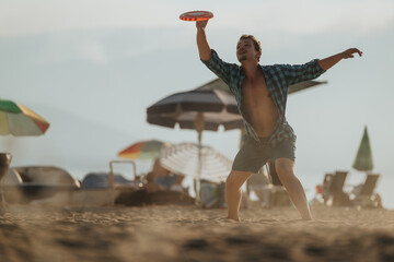 A young man enthusiastically throwing a frisbee on a sunny beach, surrounded by umbrellas and sand....