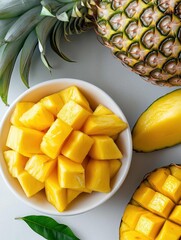 White bowl filled with diced mangoes. the mangoes are cut into small cubes and are arranged neatly in the bowl. next to the bowl, there is a whole pineapple with its green leaves still attached.