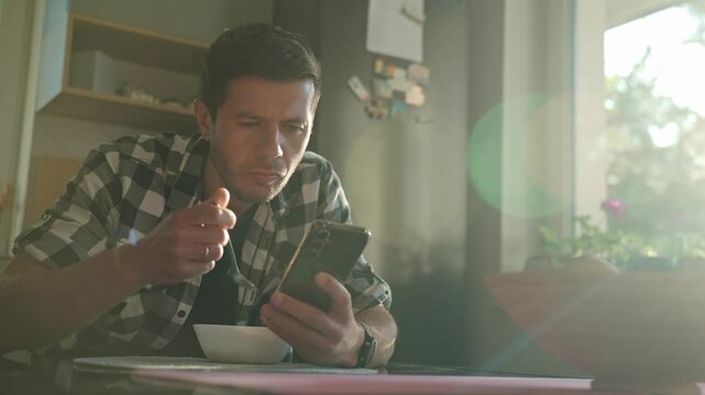 Man in plaid shirt eating oatmeal porridge from white bowl while using smartphone. Male looking at phone during breakfast. Concept of social media addiction, distraction, and digital dependency