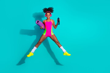 Young woman in bright sportswear jumping joyfully holding yoga mat and water bottle on a vibrant...