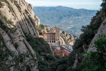 Obraz premium Views of the Monastery of Montserrat, Barcelona, Spain, from the Santa Maria gorge, surrounded by rugged rocky cliffs and dense vegetation, with distant mountains under a clear blue sky