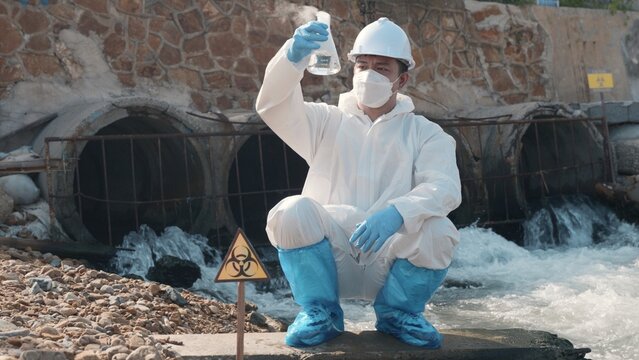 Ecologist sampling water toxic chemicals from river with test tube glass and have white smoke, Biologist wear protective suit and mask collects sample waste water from industrial, problem environment