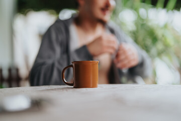 A brown mug sits on a wooden table with a blurred young male figure in the background, evoking a serene outdoor atmosphere.