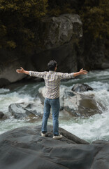 A man stands on a large rock with arms outstretched, facing a rushing river surrounded by rocks and dense forest, capturing a moment of freedom and connection with nature.