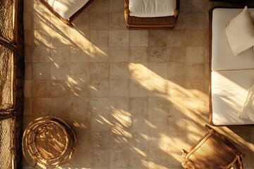 Elevated view of a sun-drenched outdoor living area with light beige tile flooring, woven furniture and white cushions  Sunlight casts shadows on the floor