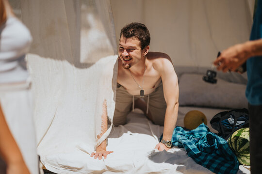 Shirtless man sitting on bedding in a sunlit outdoor setting, enjoying carefree moments and laughter. Background items add an additional summer vibe to the candid scene.