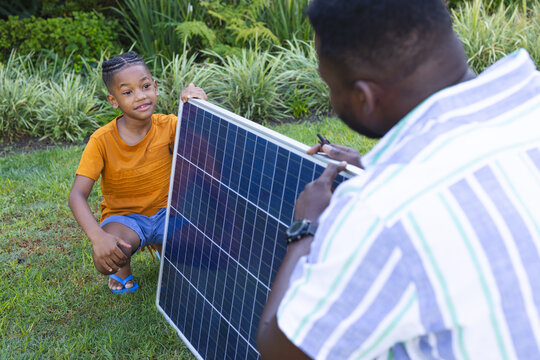 Kneeling diverse father and son installing solar panel on lawn in garden, with photovoltaic cells