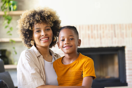 Smiling Diverse mother and son sitting on light-colored sofa in living room, with fireplace