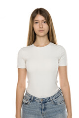Young woman with long straight brown hair wearing white fitted t-shirt and light blue jeans, neutral expression, against white background, front view portrait