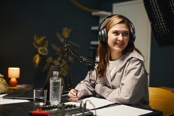 Young woman in headphones sitting at table with microphone and recording podcast in studio