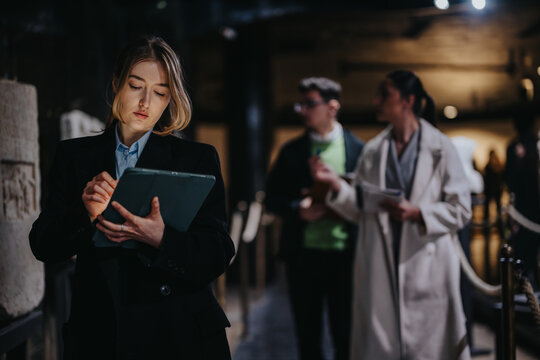 A focused woman taking notes on a tablet in a museum setting, with other visitors blurred in the background, symbolizing technology, learning, and cultural exploration in modern environments.