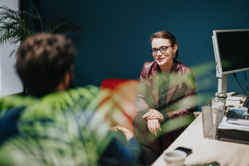 A businesswoman engages in a professional discussion with a client in a contemporary-designed office featuring a blue wall, a computer workstation, and a natural accent of green plants.