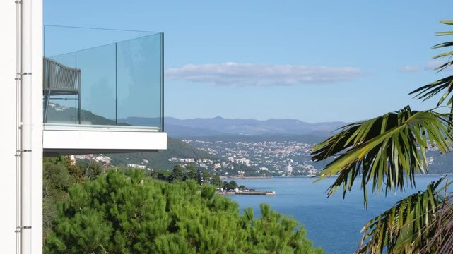 Luxury room with sea view in Lovran, Istria - Croatia. There is a palm tree in the foreground and a mountain range in the background. Seaside balcony on a sunny morning with modern glass railing.