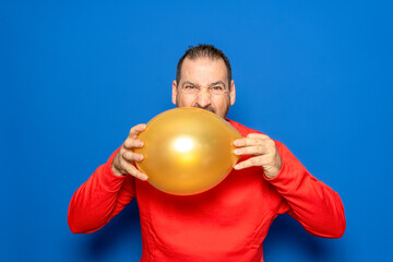 Bearded Hispanic man in his 40s wearing a red sweater holding an orange balloon annoyed and frustrated with the celebrations, isolated on blue studio background