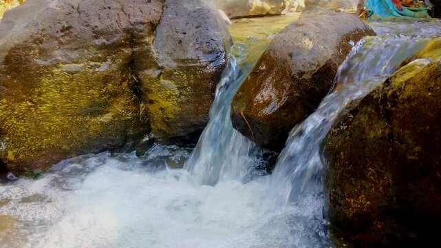 Close-up of fresh water flowing between mossy rocks, creating bubbling foam and ripples, capturing the dynamic movement and clarity of a natural stream.