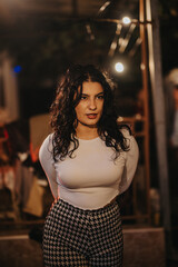 A young woman with curly hair wearing a white top and printed pants in a night setting. The warm...