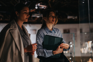 Two women in professional attire engage in discussion while exploring an art exhibit in a museum. They appear thoughtful, reflecting a moment of learning and appreciation in a cultural environment.