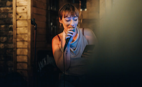 Woman comedian performing in intimate venue, engaging audience with humor. She is holding a microphone and reading from her phone, creating a lighthearted and inviting ambiance.