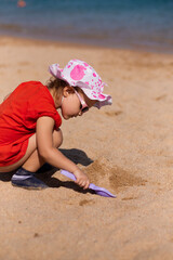 Cute kid girl in fun hat and sun glasses playing on the beach in sand with toy paddle. Builds a...