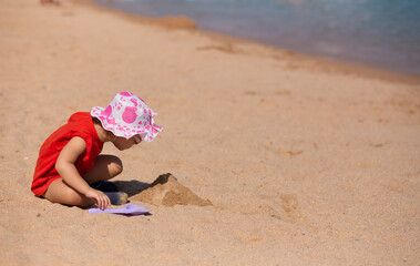 Cute baby girl in fun hat and sun glasses playing on the beach in sand with toy paddle. Builds a sand castle on sea background. Summer sunny vacations