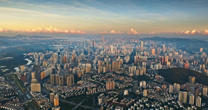 Aerial shot of the sprawling and dense urban landscape of Shenzhen featuring countless skyscrapers under a warm sunset sky near mountains.