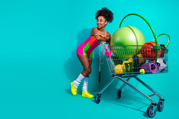 Young athletic woman in colorful activewear with shopping cart full of fitness equipment, posing...