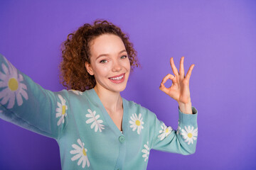 Smiling young woman with curly red hair in a stylish floral cardigan showing OK hand gesture...
