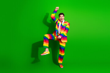 Charismatic young man in a rainbow-striped suit dances energetically against a vibrant green backdrop