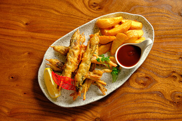 Crispy fish tempura with potato wedges, lemon, greens, and red sauce on a white plate.