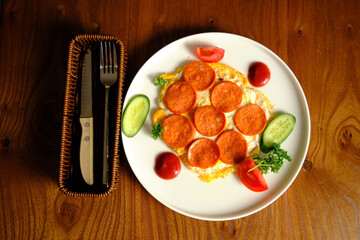 A pepperoni omelet garnished with slices of cucumber, tomato, and fresh herbs on a plate. Beside it, a woven basket holds a knife and fork on a wooden