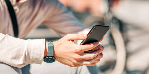Online in city. Smiling attractive guy in shirt sits on street and looks at smartphone, close up, cropped, free space