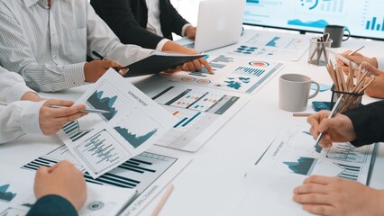 A diverse business team engages in a detailed discussion over graphs and charts displayed on paper and digital devices, fostering collaboration and strategy development. SACTR