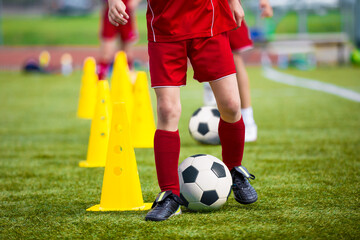 Children in red soccer uniforms practicing dribbling skills around yellow cones on a grassy field...