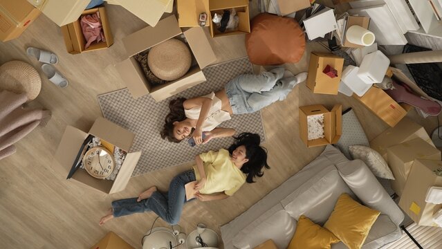 Lesbian lover looking at phone while laughing together and lying down at living room floor with cardboard box scattering. Happy couple checking at smart phone while spending time together. Myrmidon.