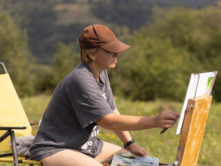 Female artist creates a landscape painting in a serene meadow surrounded by majestic mountains during a sunny day