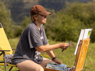 Female artist creating a painting in a tranquil meadow surrounded by majestic mountains during a sunny day