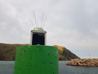 A colorful lighthouse on a green buoy overlooks a calm bay bordered by a rugged shoreline. The sky...