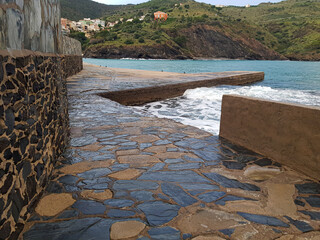 A beautiful coastal walkway lined with smooth stones, providing access to the water. Waves crash gently against the shore while the backdrop features green hills and distant homes