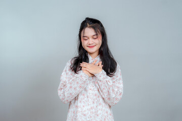 Young Asian woman with closed eyes and peaceful smile, gently placing hands over heart. Expressing...