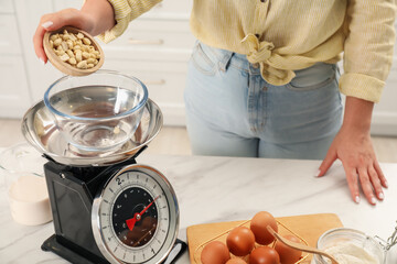 Woman adding peanuts into bowl on mechanical kitchen scale at white marble table indoors, closeup