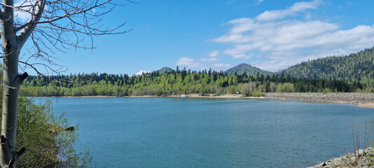 A wide shot captures the serene Lake Lepenica in Gorski Kotar, surrounded by lush green forests and distant mountains under a bright blue sky with scattered clouds