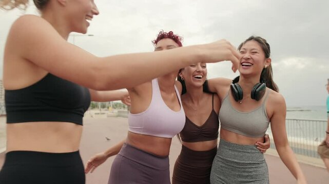 Four diverse female friends laugh and hug as they high five after a fitness workout together by the beach. They are happy and full of energy.